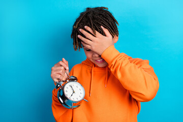 Cheerful schoolboy holding alarm clock with hand on forehead displaying a playful expression in...