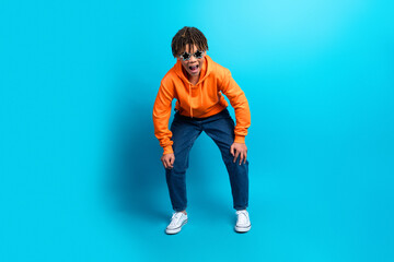 Energetic teenager dressed in orange hoodie posing with excitement on bright blue background...