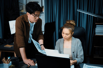 Serious face working on laptop of businesswoman with coworker giving overwork paperwork at over...