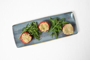 Tomatoes stuffed with vegetables and cheese, served with arugula on a plate, isolated on white background