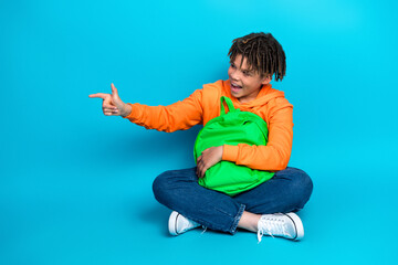 Charming young schoolboy in vibrant orange hoodie holding green backpack gesturing with joy against...