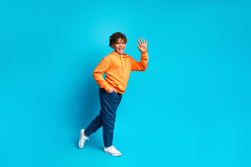 Smiling young boy in vibrant orange hoodie posing against a blue background, waving and gesturing...