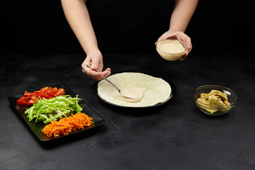 Hands of a person preparing a fresh wrap with various vegetables and sauce on a black surface, showcasing culinary skills and healthy eating concepts