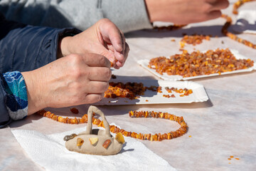 Hands crafting amber bead necklaces by stringing small amber pieces on a table with white paper plates filled with beads in a workshop