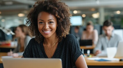 Smiling young African American businesswoman working on a laptop at her desk in a bright modern office with colleagues in the background, no logos, no brands
