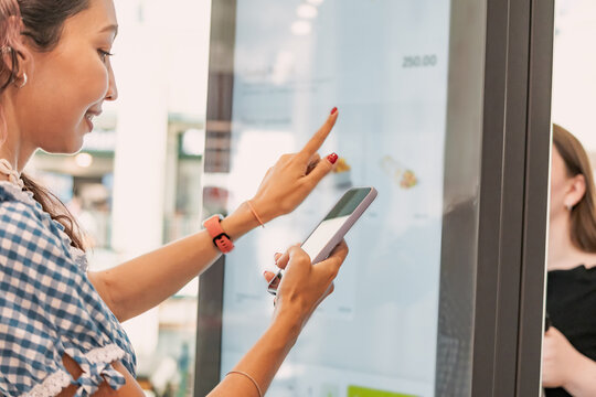 Customer using smartphone and interactive self ordering kiosk at fast food restaurant to choose meal and make contactless payment - Powered by Adobe