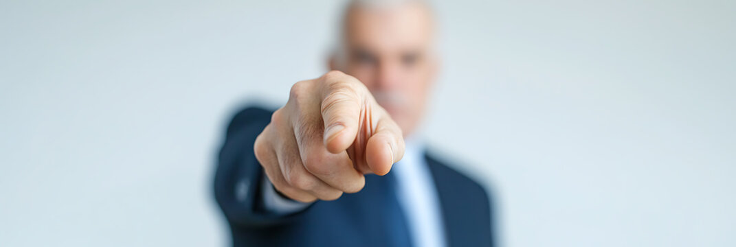 Senior man in business attire pointing directly at the viewer with a strong and direct expression, signaling a call to action or focus.