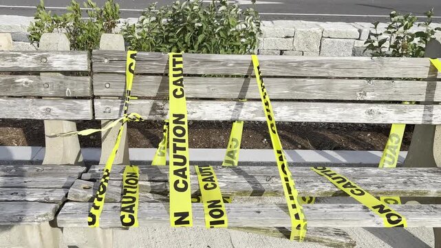 An aged wooden park bench with broken planks sits outdoors, secured with yellow caution tape marked in black text, symbolizing damage and restriction in a public setting.