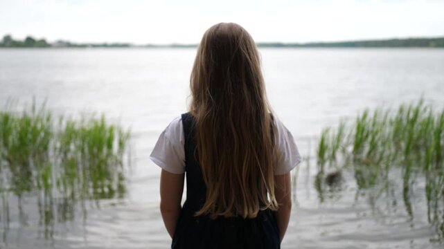 A blonde-haired girl stands on the shore of a calm lake, gazing into the distance. Peaceful outdoor scene with natural light on a quiet day. Face not visible.