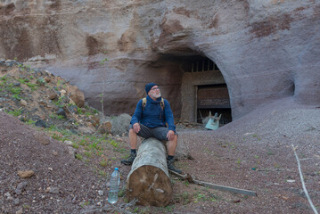 Senior Hiker Resting on a Log in Front of a Mysterious Cave Entrance