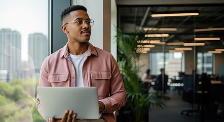 Young professional man with glasses holding a laptop and looking out a window in a modern office