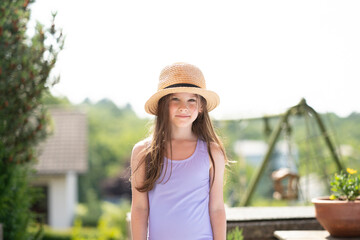 Smiling girl in a straw hat and swimsuit on a summer balcony. Concept: childhood happiness, vacation mood, sunshine joy, carefree outdoor moment.