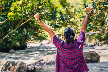 Man in plaid shirt with tied hair raising arms beside river in lush Colombian forest at countryside...