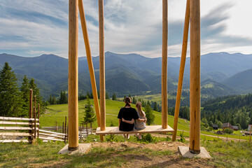 Two sisters sitting on swing with mountain view. Two young sisters sit together on a large wooden swing, hugging and enjoying a beautiful panoramic view of green forested mountains.