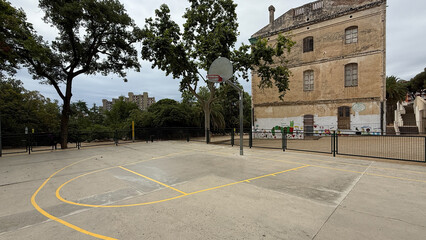 Quiet Basketball Court in Barcelona Nestled Among Lush Green Trees and an Aged Historical Building Under a Cloudy Sky