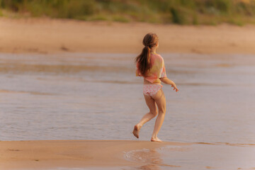 Fototapeta premium A young girl joyfully leaps into a shallow tidal pool on a beach, wearing a colorful shirt and shorts on a warm, hazy day.