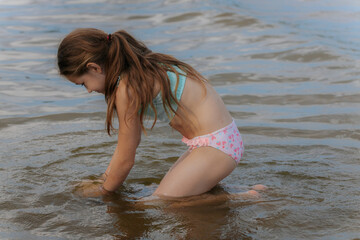 A young girl kneels in shallow water at the beach, curiously playing with the sand beneath the surface on a calm summer day.