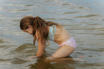 A young girl kneels in shallow water at the beach, curiously playing with the sand beneath the...