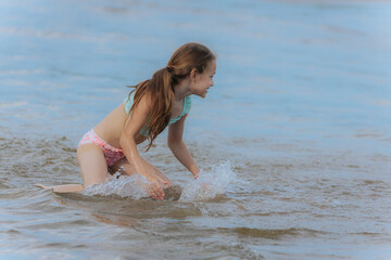 A young girl kneels in shallow water at the beach, curiously playing with the sand beneath the...