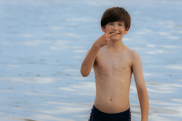 A smiling young boy stands shirtless near the water, flashing a peace sign with his fingers on a...