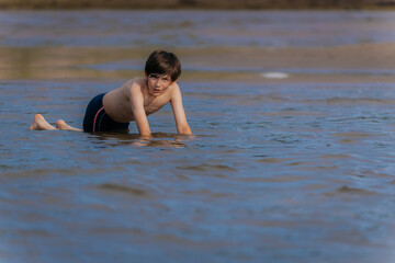 A young boy in swim trunks kneels in shallow beach water, focused and engaged in play on a sunny...