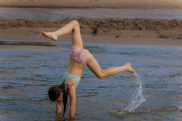 Naklejka premium A young girl performs a handstand in shallow water at the beach, showcasing strength and playfulness during a sunny day.