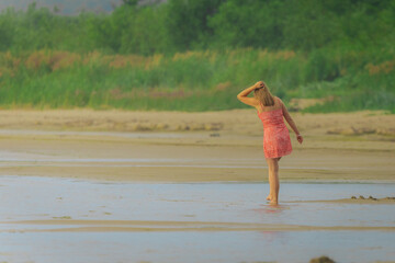 A woman in a red dress stands barefoot in shallow beach water, facing lush greenery in a peaceful...