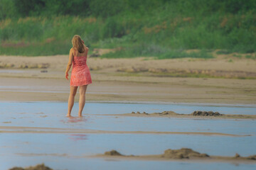 A woman in a red dress stands barefoot in shallow beach water, facing lush greenery in a peaceful...
