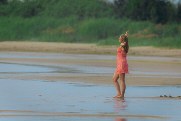 A woman in a red dress stands barefoot in shallow beach water, facing lush greenery in a peaceful...