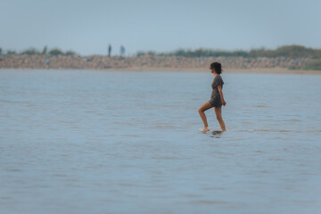 Smiling woman in a bikini runs through shallow water near a beach with tall grass in the background...