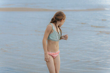 Obraz premium Young girl in a swimsuit stands in shallow beach water, looking down with a smile. Her wet hair and playful stance capture a carefree summer moment.
