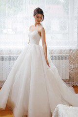 Elegant bride in flowing wedding dress poses inside sunlit room