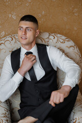 Young man in formal attire seated in a vintage chair adjusting his tie