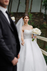 Bride in white dress with bouquet approaches groom in formal wear in a serene outdoor setting
