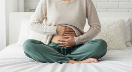 Pregnant woman in beige sweater and green pants sits on bed, hands gently cradling her belly. Image conveys serenity, anticipation, and the joy of pregnancy.
