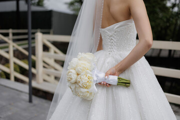 Bride in elegant wedding dress holding bouquet of peonies on a sunny day outdoors