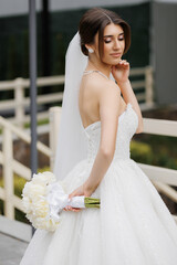 Beautiful bride in elegant white gown holding bouquet outdoors during wedding preparations