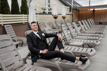 Groom relaxing in a suit by the poolside loungers at a summer wedding event