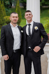 Two friends dressed in formal suits pose together outdoors during a wedding celebration