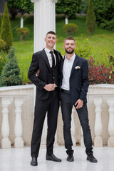 Two friends pose happily at an outdoor venue wearing formal suits during a sunny day