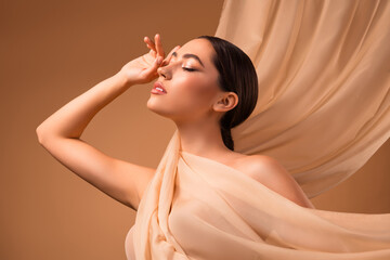 Portrait of a young brunette woman against a beige background wrapped in soft fabric with elegant...