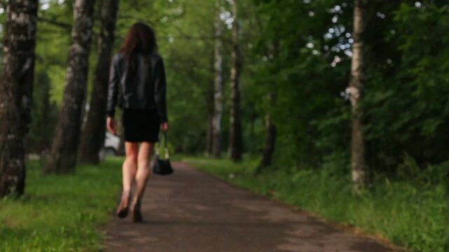 Business slim woman with sexy legs walking on a road in high heels fashion shoes, leather jacket in summer day among green trees. Attractive success lady moving in fashion style dress. Back view