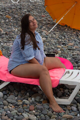 Vertical travel photo. Young adult white fat woman with bare legs, shirt with stripe sits on plastic sun chaise lounge on pebble sea beach. Sunny day. Tourism, summer rest, trip, relax, resort