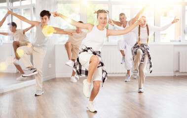Young girl in bright clothes, together with group of teenage friends, learn to dance hip hop in choreographic hall. Students move to modern music, do moonwalking, squats, bends, work with body