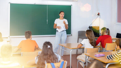 Young woman teacher explaining subject to schoolgirls and schoolboys during lesson in school.