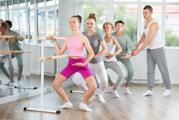 Young teen student of ballet school near ballet bare performs plie under guidance of experienced...