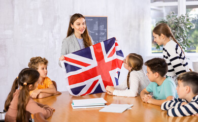 Decent teacher showing flag of Great Britain to group of preteen schoolchildren in classroom during...
