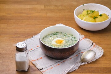 Cold sorrel soup with boiled egg and greens in a brown ceramic bowl on a wooden table. Served with boiled potatoes. Wild greens dishes.