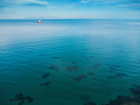 Aerial view of a red cargo ship navigating on a calm blue sea with visible seabed and distant coast on the horizon