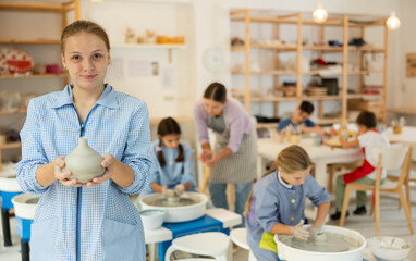 Teenage holds a clay product against the background of a teacher with children. Children working at...